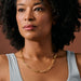 A close-up of a woman wearing a gold chain link necklace and a light blue top, set against a solid warm brown background.