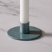 A close-up of a round blue/gray metal candle holder base with a matching teal cylindrical holder, holding a white candle on a light marble surface.