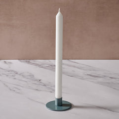 A tall white taper candle in a round blue/gray metal holder, set on a white marble surface against a textured beige-brown background.