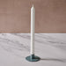 A tall white taper candle in a round blue/gray metal holder, set on a white marble surface against a textured beige-brown background.