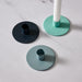 Three round metal candlestick holders in blue, blue/gray, and turquoise, with one holder shown holding a white candle on a light marble surface.