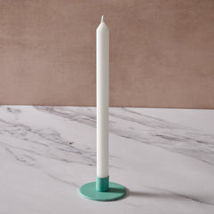A white taper candle in a round turquoise holder, set on a white marble surface with a textured beige background.