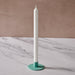 A white taper candle in a round turquoise holder, set on a white marble surface with a textured beige background.