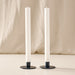 Two tall natural white taper candles in round blue metal holders, standing on a light surface against a soft beige fabric backdrop.