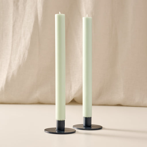 Two tall, slim pillar candles in a sage green color, standing in blue metal candle holders with round bases against a soft beige fabric backdrop.