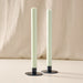 Two tall, slim pillar candles in a sage green color, standing in blue metal candle holders with round bases against a soft beige fabric backdrop.