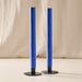 Two tall cobalt blue taper candles in blue metal holders, standing upright against a soft beige fabric backdrop.