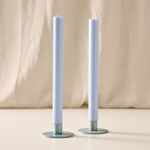 Two tall mint taper candles in round blue/gray holders, standing on a light beige surface with a draped beige fabric backdrop.