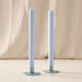 Two tall mint taper candles in round blue/gray holders, standing on a light beige surface with a draped beige fabric backdrop.