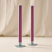Two slim blackberry dinner candles in round blue/gray metal holders, standing on a light surface against a draped cream fabric backdrop.