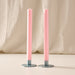 Two tall ginger pink colored taper candles standing upright in round blue/gray metal holders against a soft beige fabric backdrop.