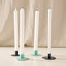 Four taper candles in white holders with round turquoise and blue bases, arranged on a light surface against a soft beige fabric backdrop.
