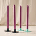 Four taper candles in a blackberry color, each set in a round candle holder base with two different colors: blue and turquoise. The candles are arranged on a light beige surface against a draped cream fabric background.