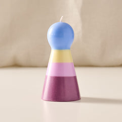 A pawn shaped candle with a rounded blue top and a tapered base in horizontal pastel stripes of yellow, pink, and purple, standing on a light beige surface against a soft cream background.