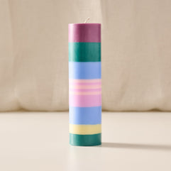 A tall cylindrical candle with horizontal pastel stripes in purple, green, blue, pink, and yellow, standing upright on a light surface against a soft beige fabric background.