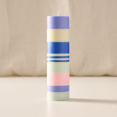 A tall, cylindrical striped candle with pastel bands of purple, cream, blue, white, mint green, and pink, standing upright on a light beige surface against a soft cream fabric backdrop.