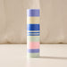 A tall, cylindrical striped candle with pastel bands of purple, cream, blue, white, mint green, and pink, standing upright on a light beige surface against a soft cream fabric backdrop.
