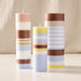 Three striped pillar candles in pastel shades of pink, blue, yellow, and brown, standing on a white surface against a soft beige fabric backdrop.