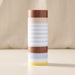 A cylindrical pillar candle with horizontal stripes in brown, cream, light blue, and pale yellow, standing upright on a light surface against a soft beige fabric background.