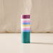 A cylindrical pillar candle with horizontal pastel stripes in purple, white, pink, light blue, yellow, and teal, standing upright on a light beige surface against a soft beige fabric backdrop.