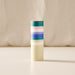 A tall, cylindrical candle with horizontal stripes in teal, pink, white, and blue, with the lower half in solid cream-colored wax, set against a soft beige fabric backdrop.