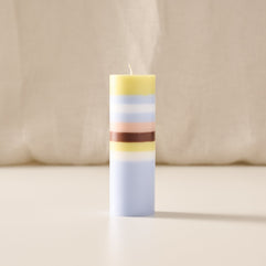 A tall, cylindrical striped candle with pastel yellow, light blue, white, peach, and dark brown bands, standing upright on a light beige surface against a soft beige fabric backdrop.