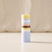 A tall, cylindrical striped candle with pastel yellow, light blue, white, peach, and dark brown bands, standing upright on a light beige surface against a soft beige fabric backdrop.
