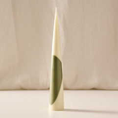 A tall, tapered cream-colored candle stands upright on a light surface against a soft beige fabric backdrop. The candle features an abstract olive-green curved shape wrapping around its lower half. Warm, diffused lighting creates a minimalist, sculptural appearance with a subtle shadow cast to the right.
