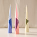 Three tapered decorative candles with geometric color-blocked patterns, standing upright on a light surface against a soft beige fabric backdrop. The candles are light blue and navy, pink with purple and peach sections, and cream with olive green sections.
