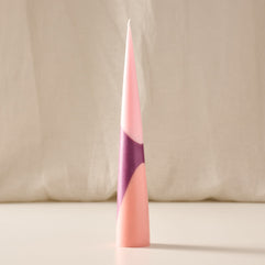 A tall, slender cone-shaped candle with a geometric color-blocked design in shades of pink, peach, and purple, standing upright on a light surface against a soft beige fabric backdrop.