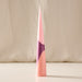 A tall, slender cone-shaped candle with a geometric color-blocked design in shades of pink, peach, and purple, standing upright on a light surface against a soft beige fabric backdrop.
